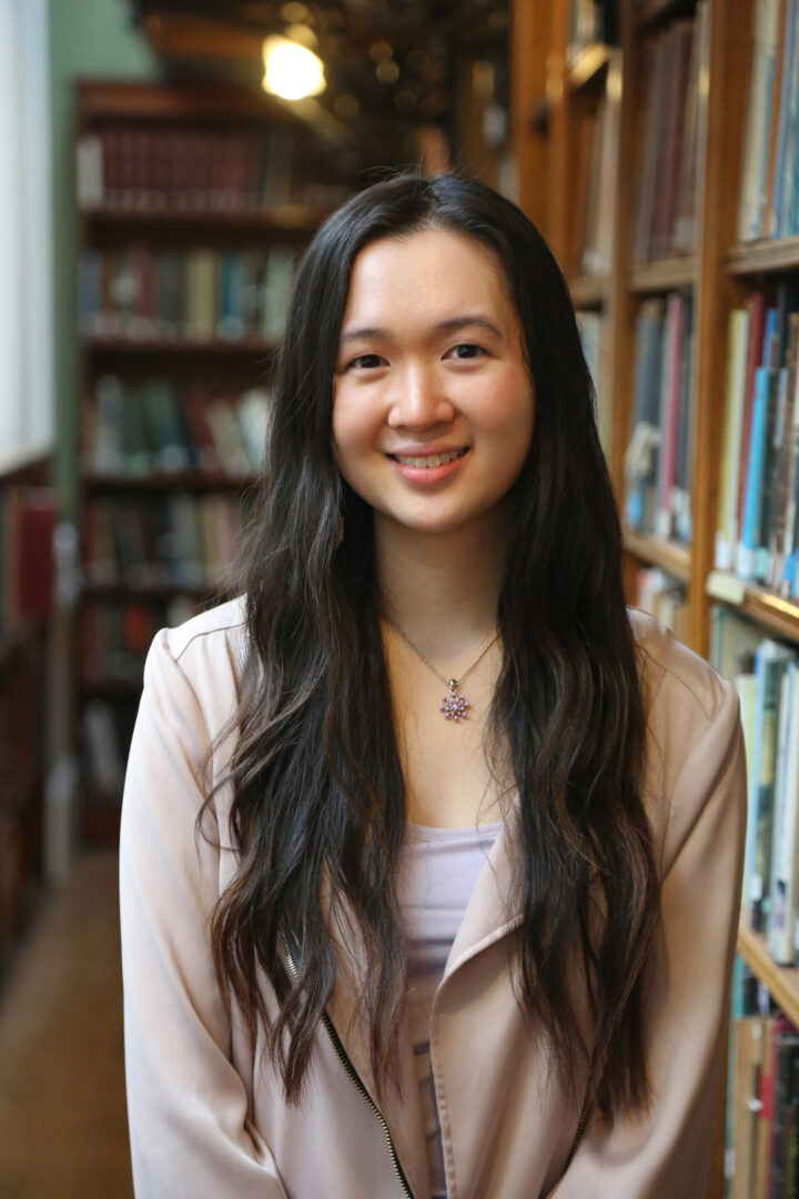 Katie smiles at the camera, standing in front of a bookshelf wearing a pink blazer.