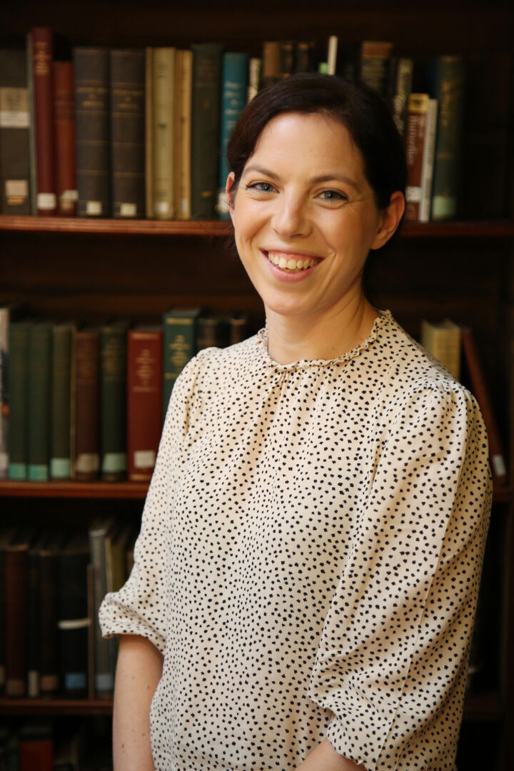The head of engagement team stands smiling in front of a bookcase in a white patterned shirt.