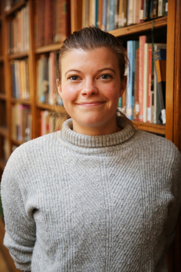 Ayesha smiles at the camera, wearing a grey jumper in front of a bookshelf. Her hair is tied back into a ponytail.