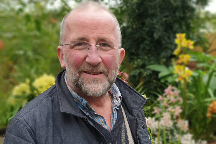 A man with a beard and glasses in a dark coat stands in a botanic garden