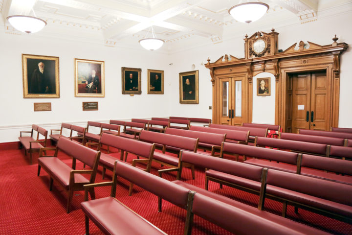 An image of a large meeting room, with bench-style seating arranged in rows. There are two sets of double doors at the back of the room, and portraits of famous naturalists (including Darwin's) on the walls.