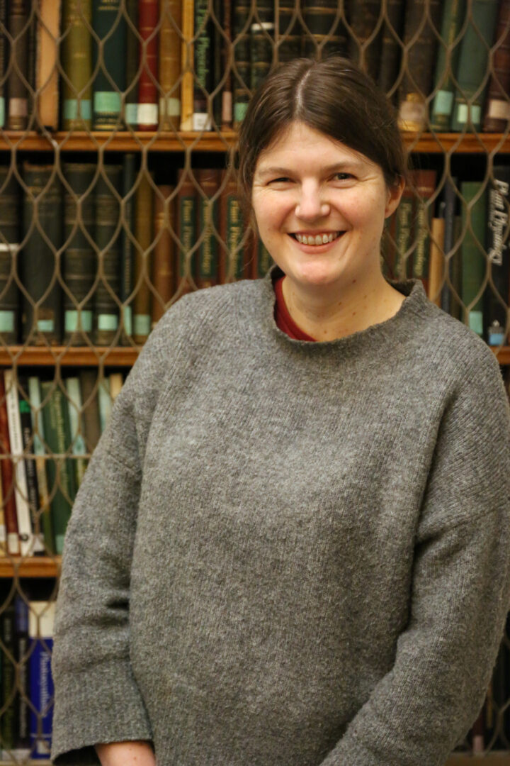 Alice smiles facing the camera, wearing a grey woolly jumper whilst stood in front of a bookshelf.