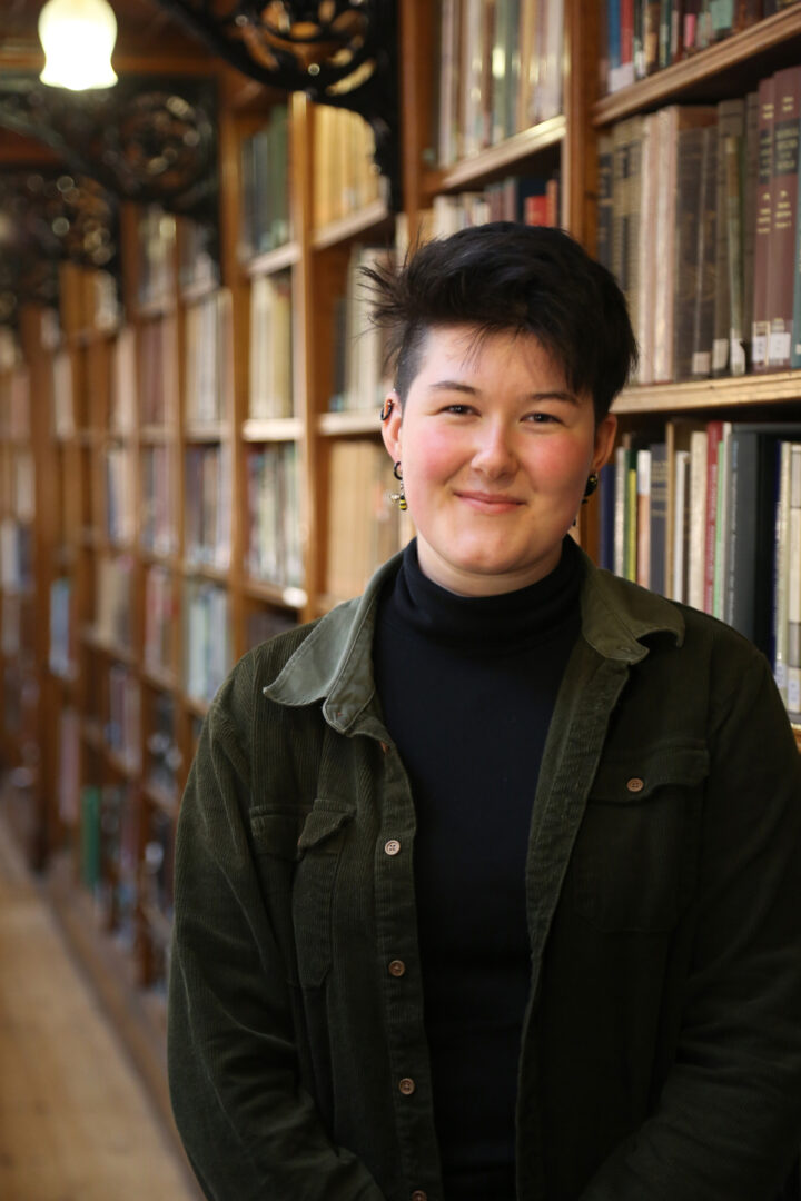 Scarlet stands smiling in front of a bookcase, wearing a green shirt over a black top.