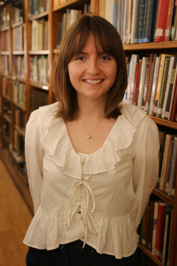 Georgia stands facing the camera, her hands behind her back, as she smiles in front on the bookshelf. She is wearing a white frilly top.