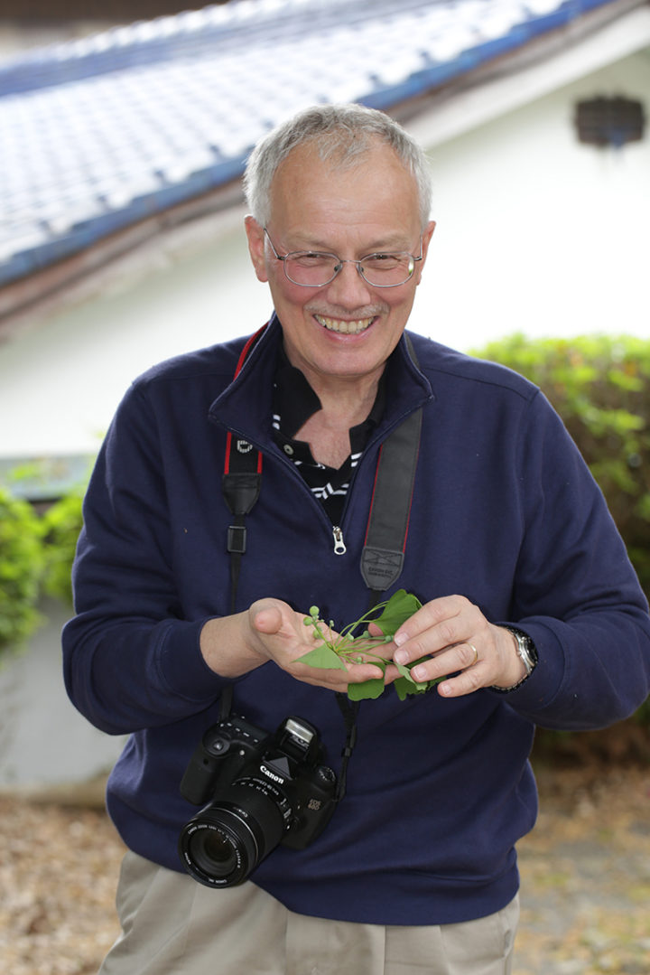 Professor Peter Crane smiles and holds a plant in his hand, with a camera round his neck