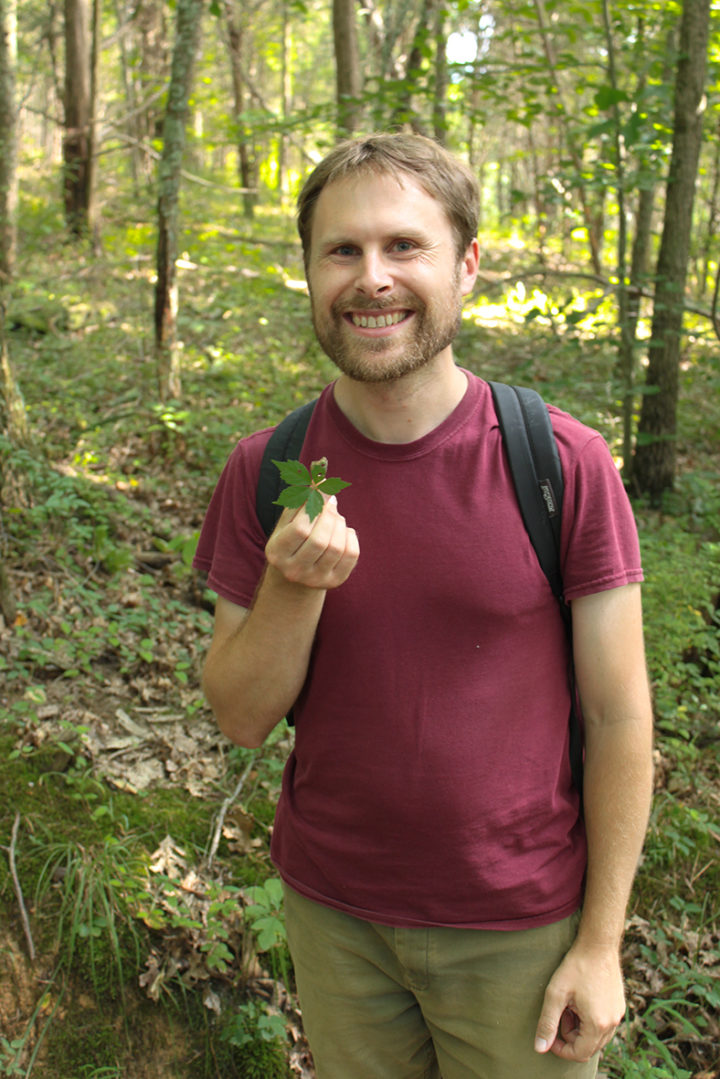 Charley Eiseman stands in a woodland wearing a backpack holding some leaves and smiling