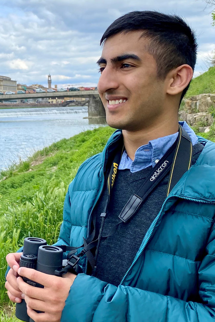 Kabir Kaul stands on a grassy riverbank in a blue puffer jacket holding binoculars
