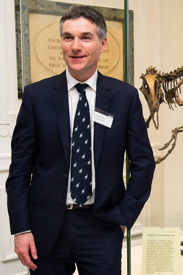 Professor Paul Upchurch stands in a suit in front of a dinosaur skeleton