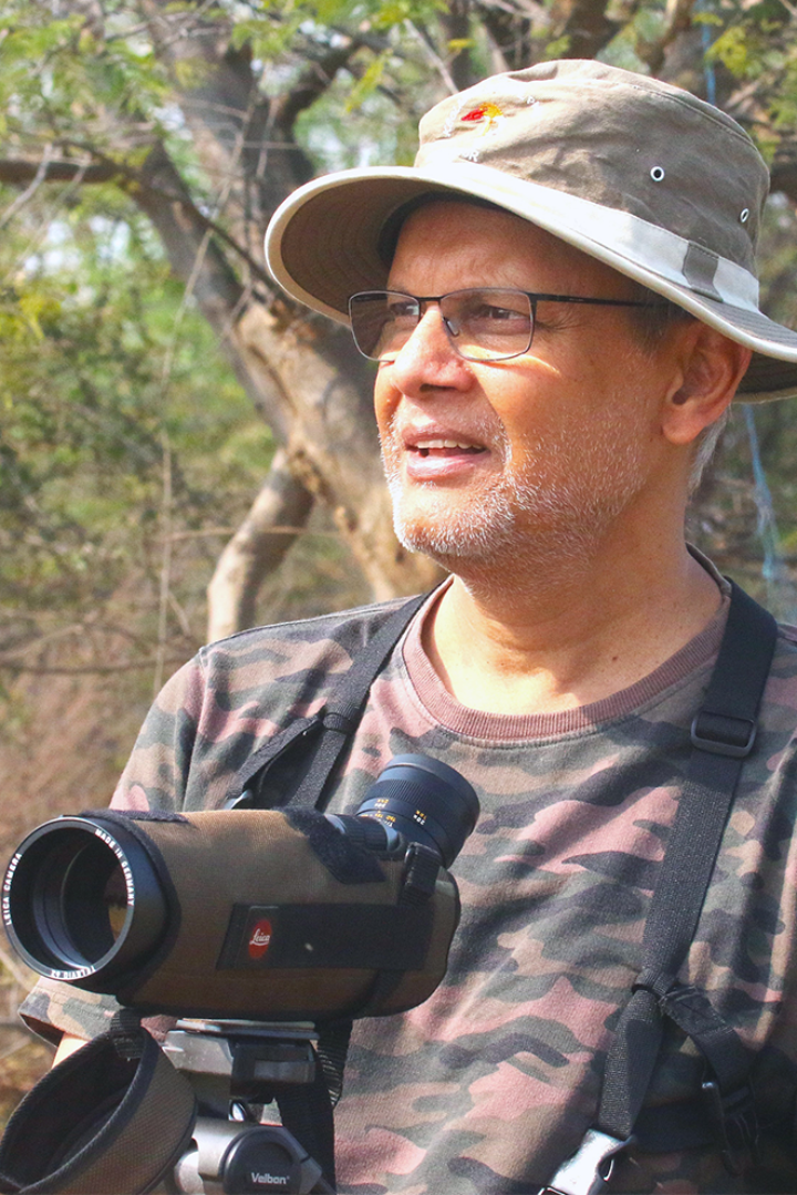 Man in hat, glasses and mottled camouflage T-shirt holds binoculars in front of tree, looking into distance