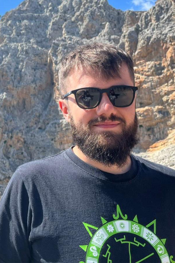 Bearded man with dark hair stands in front of mountain wearing sunglasses