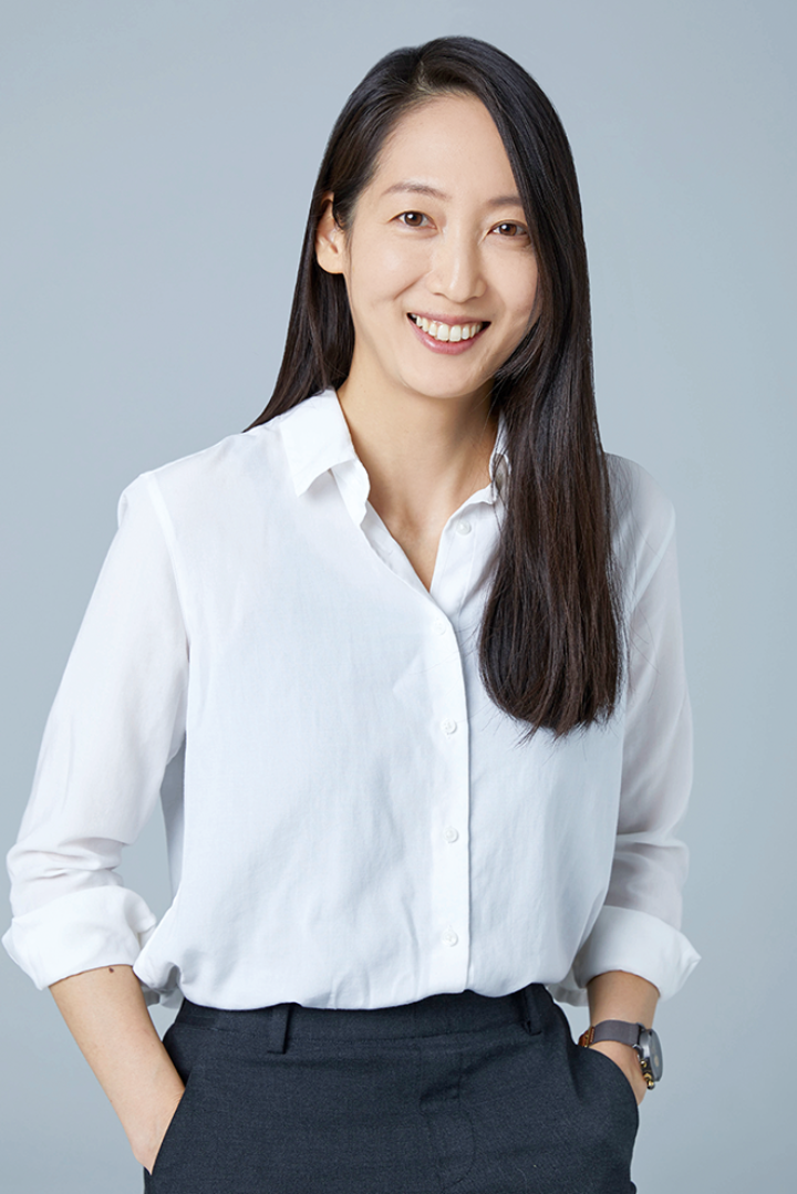 Woman in white shirt and black trousers stands with hands in pockets, long dark hair over one shoulder