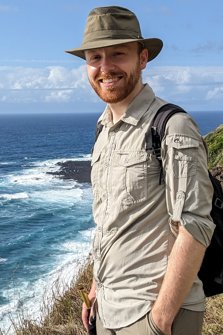 Bearded man wearing hat and khaki clothes stands on coastline, blue sky behind him