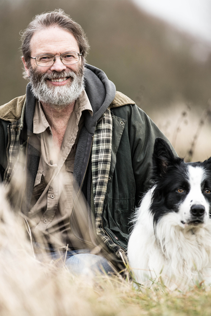 Bearded man sits in field smiling at camera, black and white dog beside him