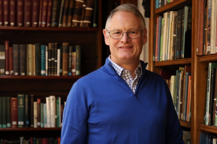 A man with glasses in a blue jumper stands in front of shelves of old books