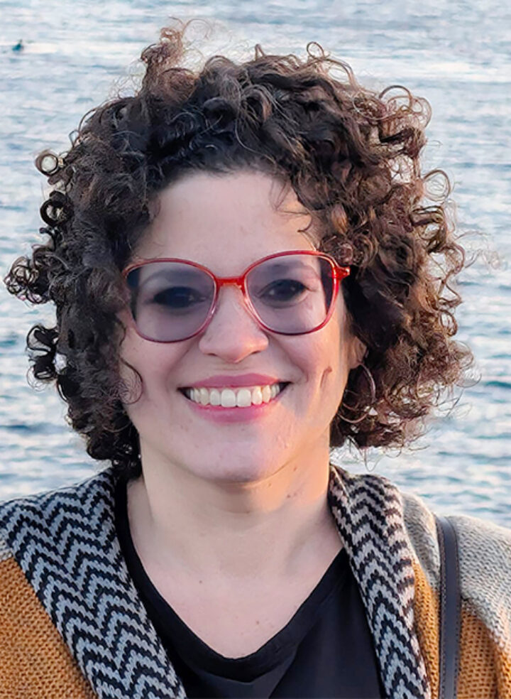 A woman with red-framed glasses and mustard yellow top stands and smiles with her brown curly hair blowing in the breeze