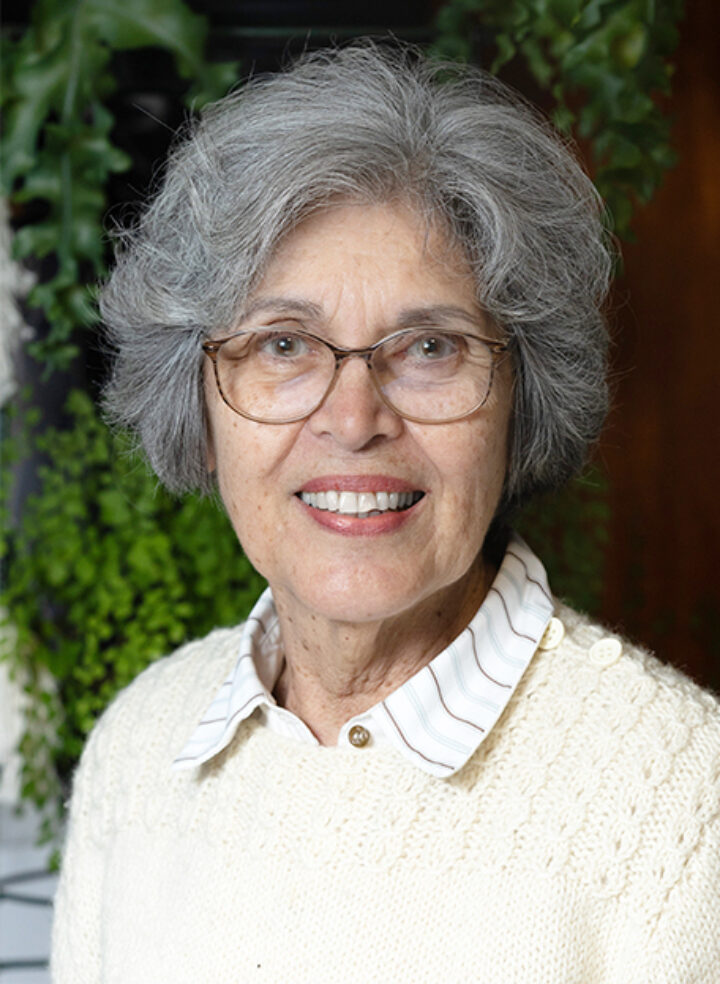 A smiling woman with chin length salt and pepper hair and glasses poses wearing a cream-coloured knitted sweater