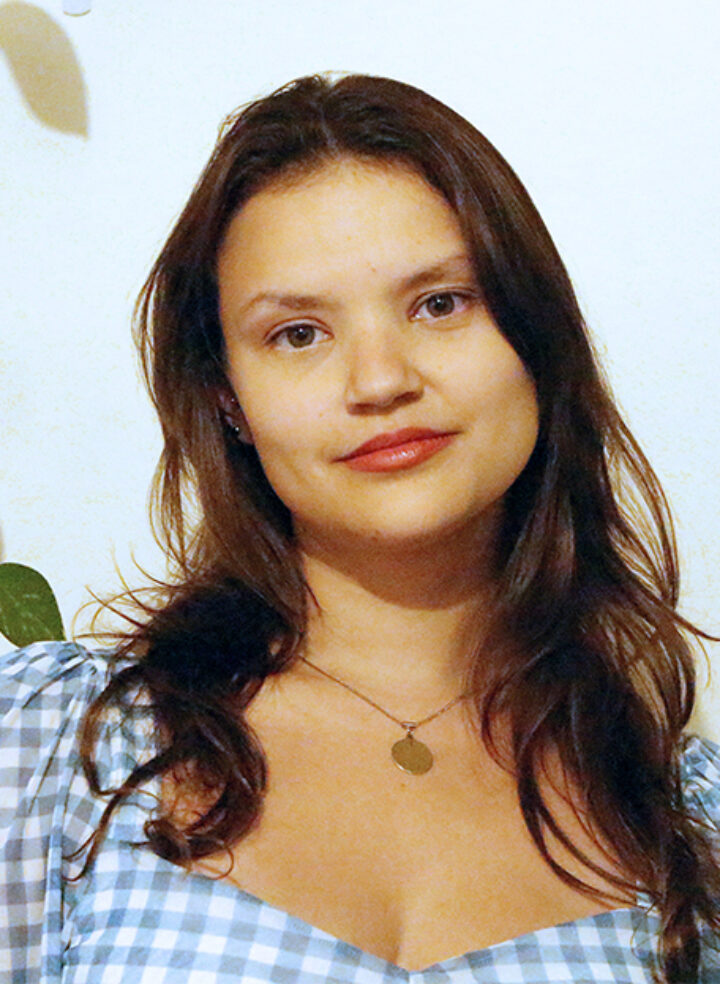 A young woman in a blue and white gingham top and long brown hair stands in front of a pale wall