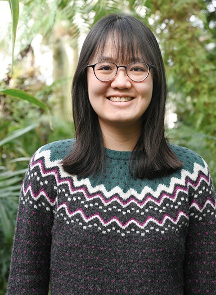 Smiling woman with glasses in a green sweater and shoulder length dark hair stands in amongst tall plants