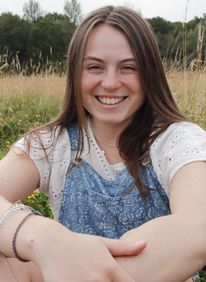 A woman with long brown hair sits down in a field, in a white T-shirt and dungarees, smiling with her arms crossed