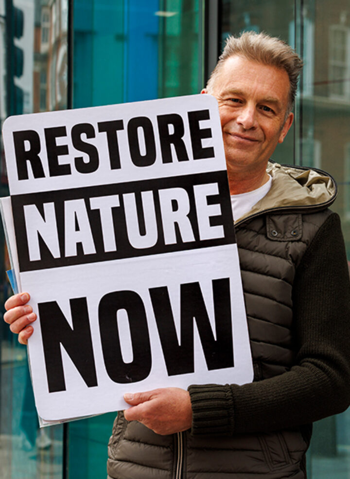 A man with short light hair in a puffer jacket smiles and holds a sign saying 'Restore nature now'