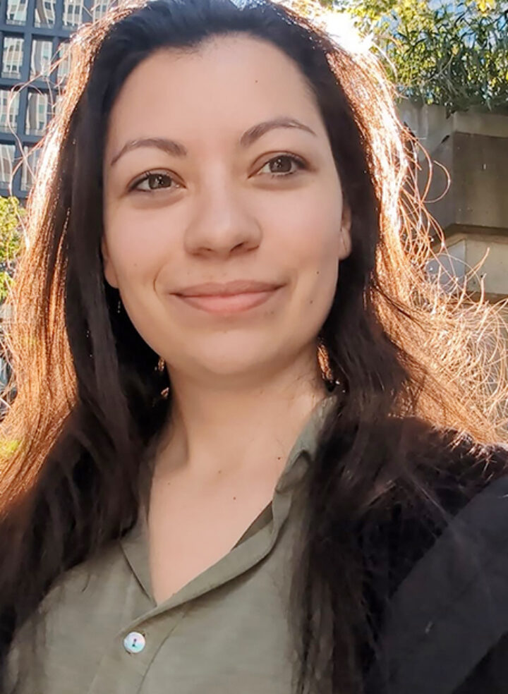 A young woman with long brown hair stands with sunlight behind her and smiles