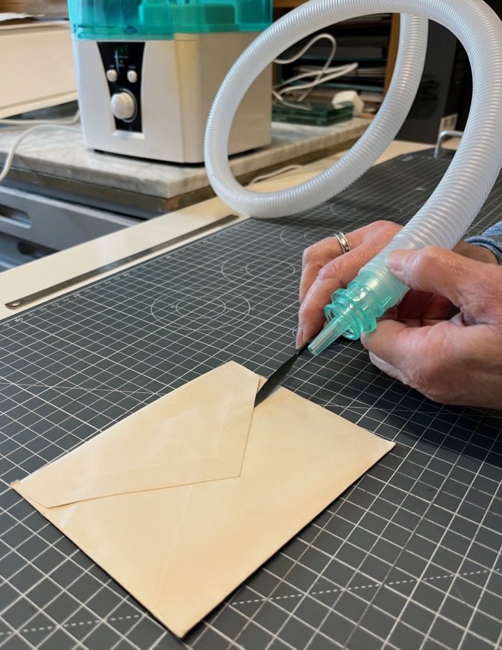 The conservator uses a nozzle to direct damp air at the seal edge of an envelope while using a thin spatula to tease the envelope open