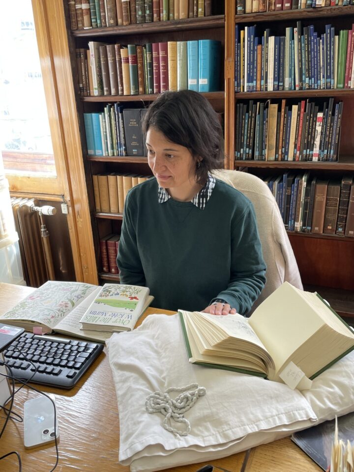 An image of a young woman sat at a computer, with a book open on a book support beside her.