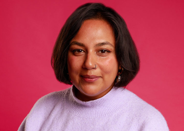 A woman with dark hair sits in front of red background in a light coloured top