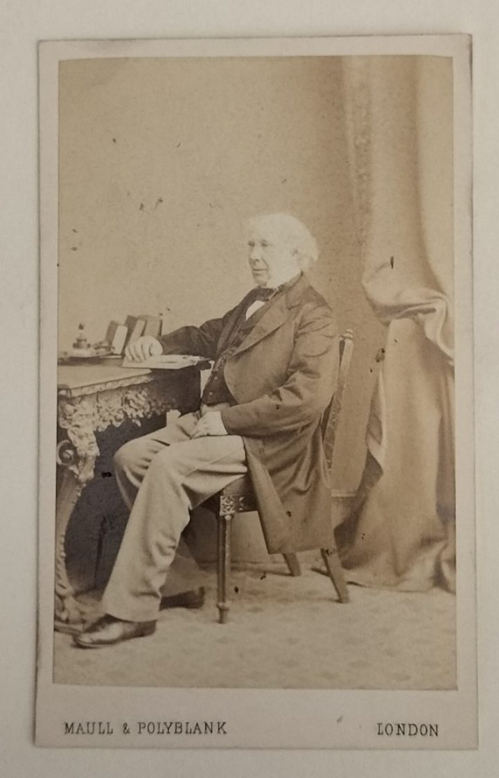 A sepia photograph of a man sitting in a chair with his arm resting on a book on the table.