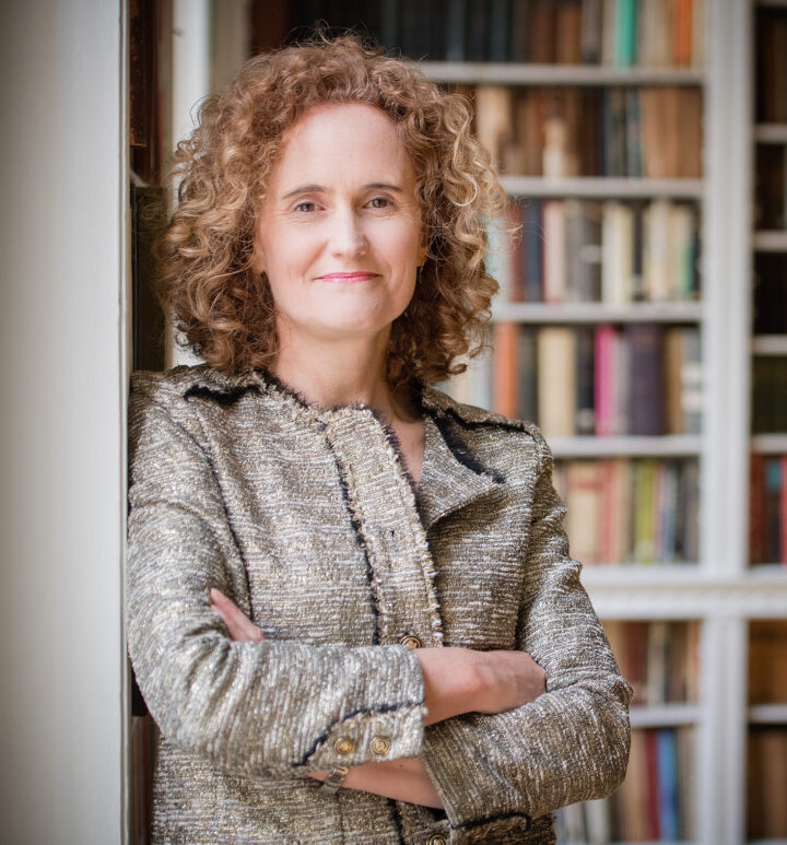 The CEO of the Linnean Society leans smiling against the wall in the library, wearing a cream jacket.