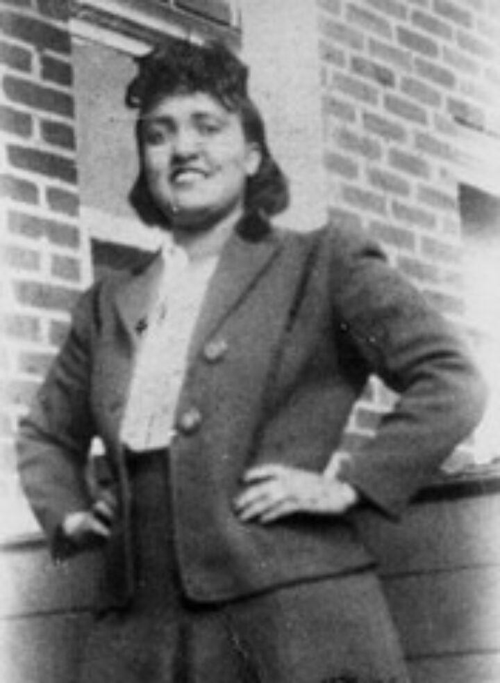 A black and white photograph shows Henrietta Lacks looking directly at the camera and smiling. She has her hands on her waist, wearing a twede skirt suit.