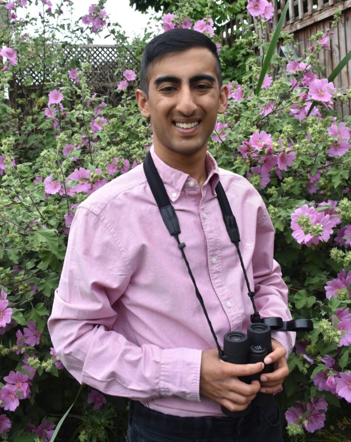 Kabir Kaul smiling, holding a pair of binoculars, in front of a flowering shrub