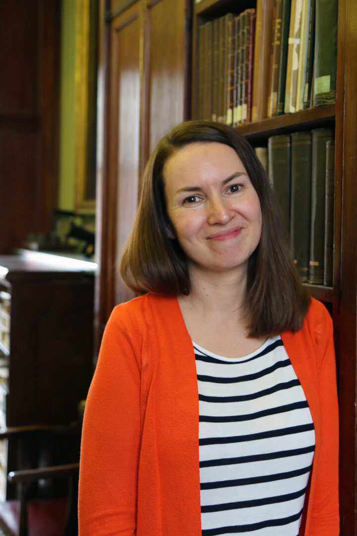 The publications manager stands smiling in an orange cardigan and striped t-shirt in front of a book case.