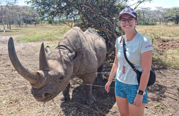 A woman, standing on the right, stands next to a rhinoceros on the left in a savannah setting