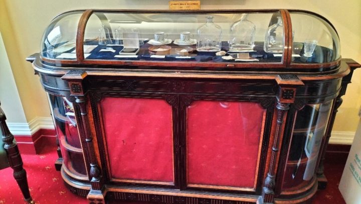 A dark wooden, oval shaped cabinet. It is about one and a half meters across with a curved glass case at the top. The front two doors in the base have been covered with red tissue paper on the inside and small items of crockery and medals can be seen under the glass top.